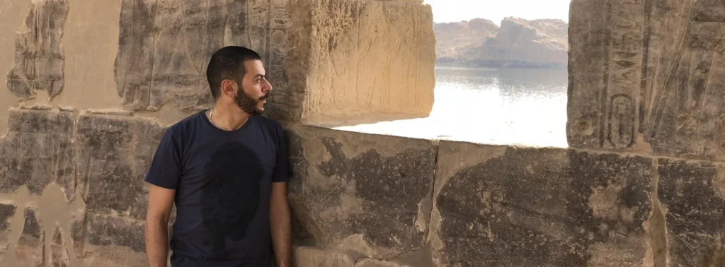 Paulo Farah encostado em um muro de pedra com inscrições antigas, olhando para o rio ao fundo