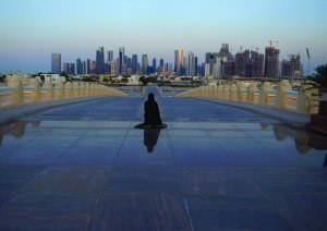 Vista do horizonte de arranha-céus de Doha ao entardecer, com uma figura solitária sentada no meio da esplanada