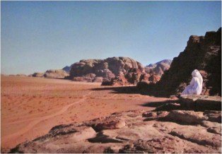 Deserto de areia avermelhada com formações rochosas ao fundo e uma figura de véu branco sentada sobre a pedra