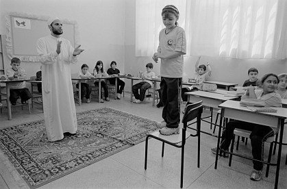 Fotografia em preto e branco de uma sala de aula: um homem de túnica branca fala em pé sobre um tapete de oração, diante de crianças nas carteiras